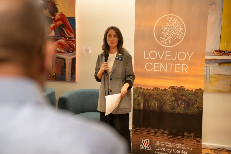 A woman stands next to a retractable banner and speaks into a microphone.